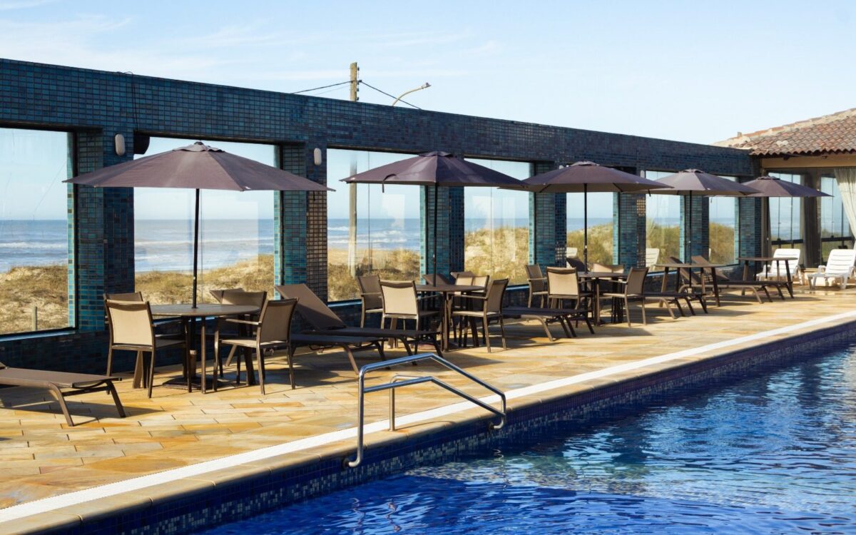 Piscina com borda azulada em primeiro plano, deck de madeira, cadeiras brancas sob guarda-sóis marrom, edifício de tijolos azuis, dunes e mar ao fundo