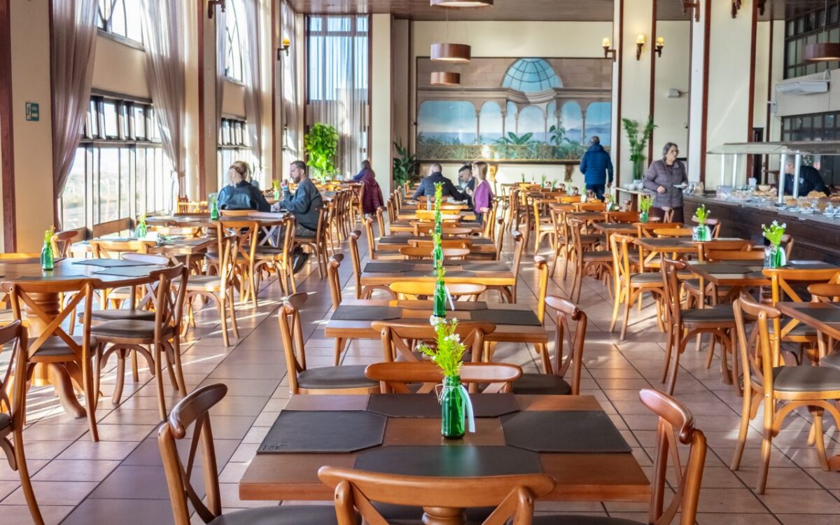 Interior de restaurante com mesas de madeira alinhadas, cadeiras estofadas, janelas amplas, teto metálico, luminária pendente e pessoas ao fundo