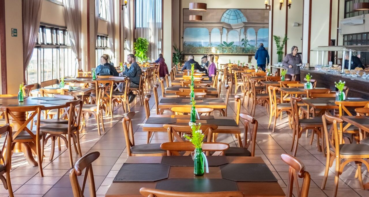 Interior de restaurante com mesas de madeira alinhadas, cadeiras estofadas, janelas amplas, teto metálico, luminária pendente e pessoas ao fundo