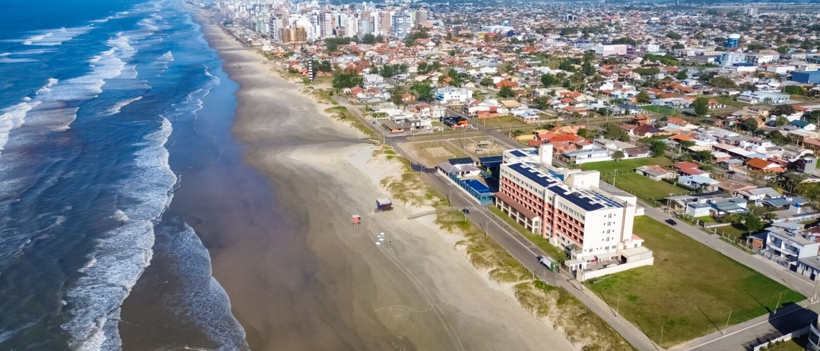 Vista aérea de praia com ondas, areia, edifícios à beira-mar, residências e montanhas ao fundo sob céu azul claro