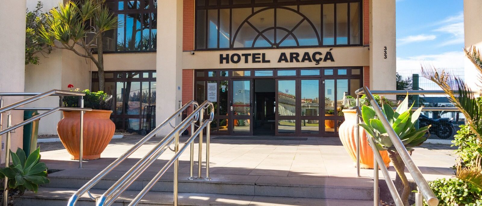 Entrada de um hotel com a palavra "ARAÇÁ" escrita na fachada, rampas metálicas, vasos de cerâmica laranja, plantas decorativas e vista para o mar ao fundo