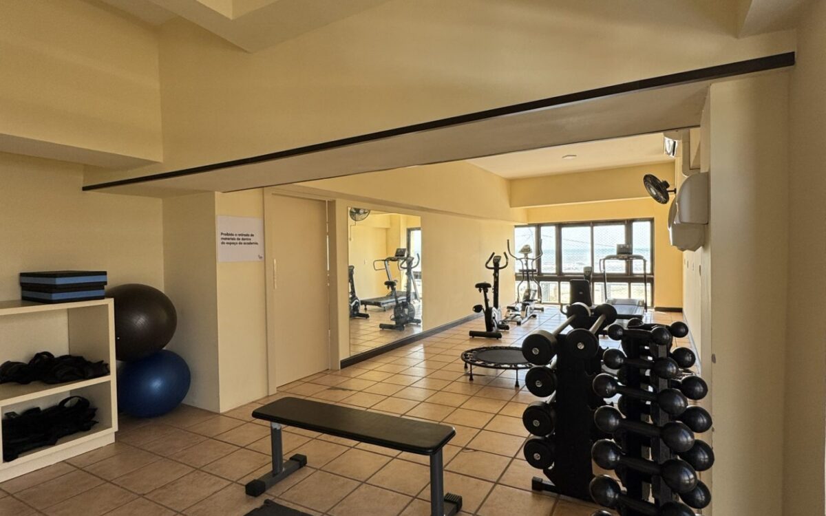 Espaço de exercícios com halteres organizados em rack, banco de musculação, bolas de ginástica, espelhos e equipamentos de cardio ao fundo