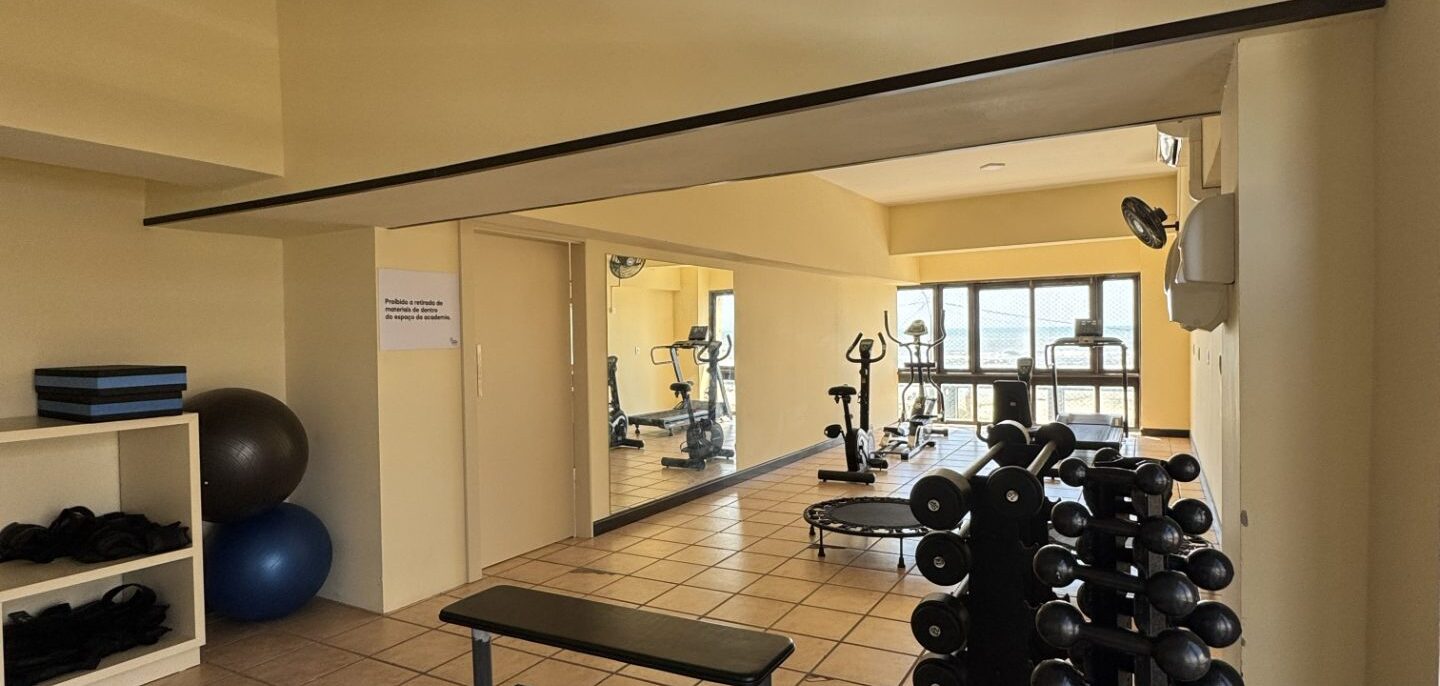 Espaço de exercícios com halteres organizados em rack, banco de musculação, bolas de ginástica, espelhos e equipamentos de cardio ao fundo