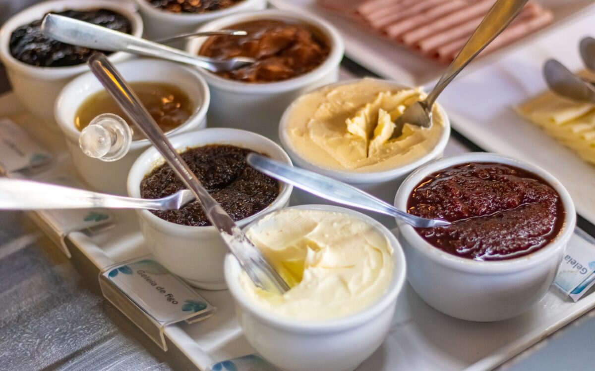 Diversos potes brancos com colheres contendo geleias, cremes e pastas de cores variadas dispostos sobre uma superfície branca