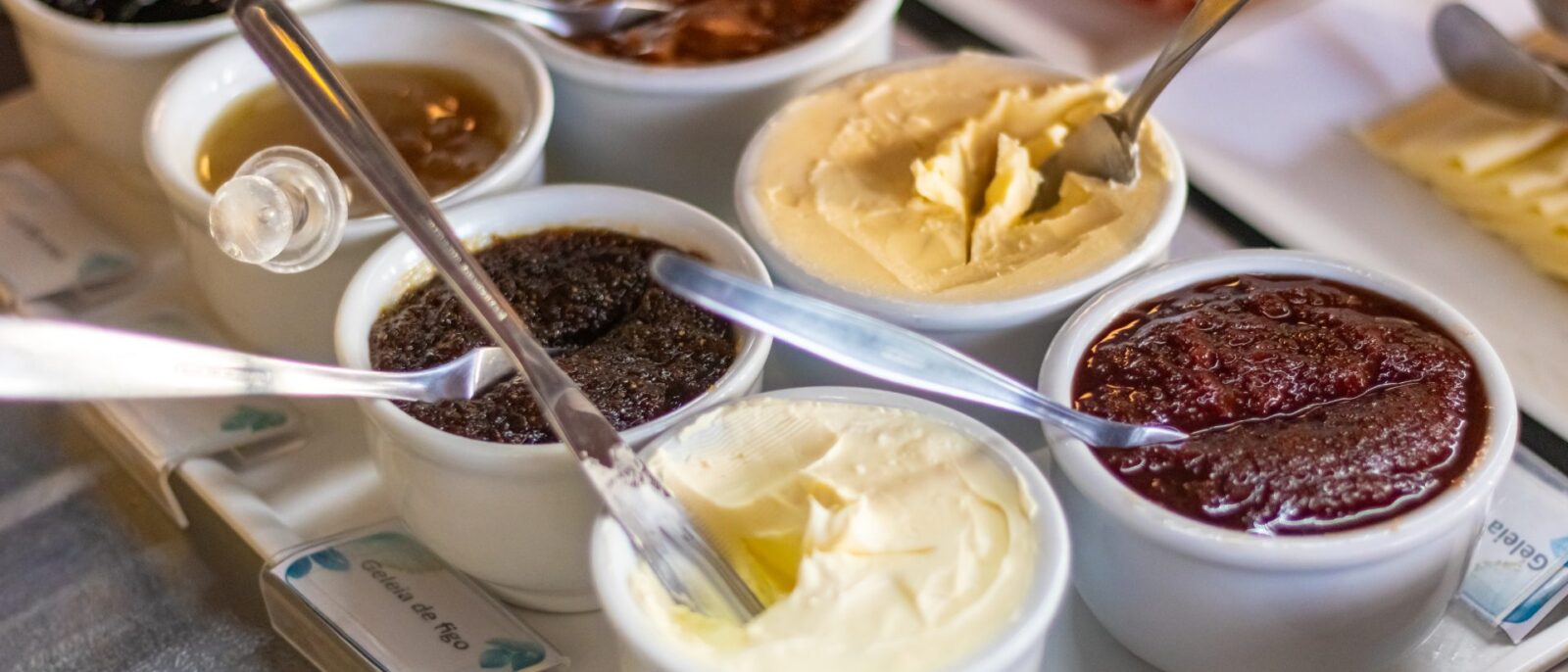 Diversos potes brancos com colheres contendo geleias, cremes e pastas de cores variadas dispostos sobre uma superfície branca