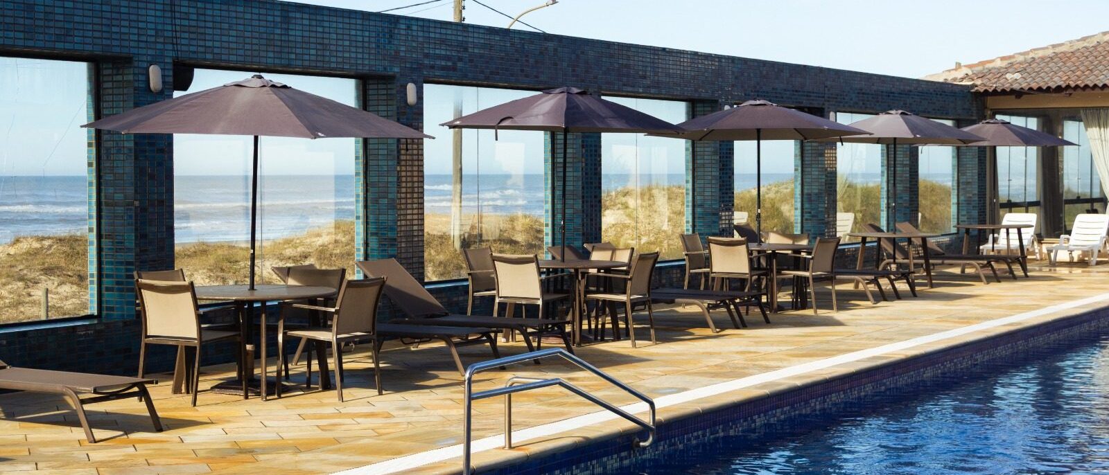 Piscina com borda azulada em primeiro plano, deck de madeira, cadeiras brancas sob guarda-sóis marrom, edifício de tijolos azuis, dunes e mar ao fundo