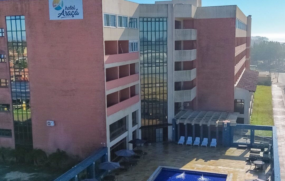 Vista aérea de um hotel com piscina retangular azul, espreguiçadeiras com guarda-sóis, edifícios de alvenaria vermelha e cinza, sob céu azul com nuvens