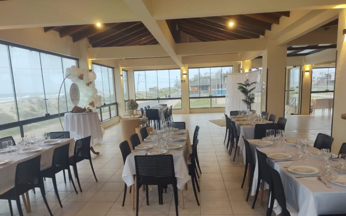 Salão de eventos com mesas compridas, cadeiras pretas, decoração floral branca, janelas amplas com vista para paisagem, iluminação moderna e teto com acabamento
