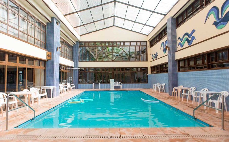 Piscina coberta com água azul-turquesa, cercada por cadeiras brancas, pisos de madeira, colunas azuis, paredes bege com detalhes em azul e teto com claraboias