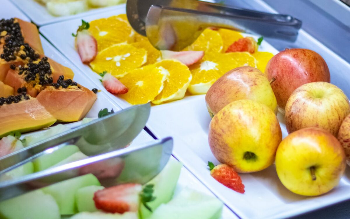 Arranjo de frutas e alimentos em pratos brancos com maçãs, laranja, melão, morango, frutas com sementes pretas e sanduíches com talheres