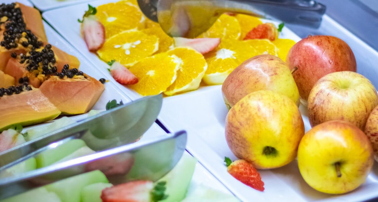 Arranjo de frutas e alimentos em pratos brancos com maçãs, laranja, melão, morango, frutas com sementes pretas e sanduíches com talheres