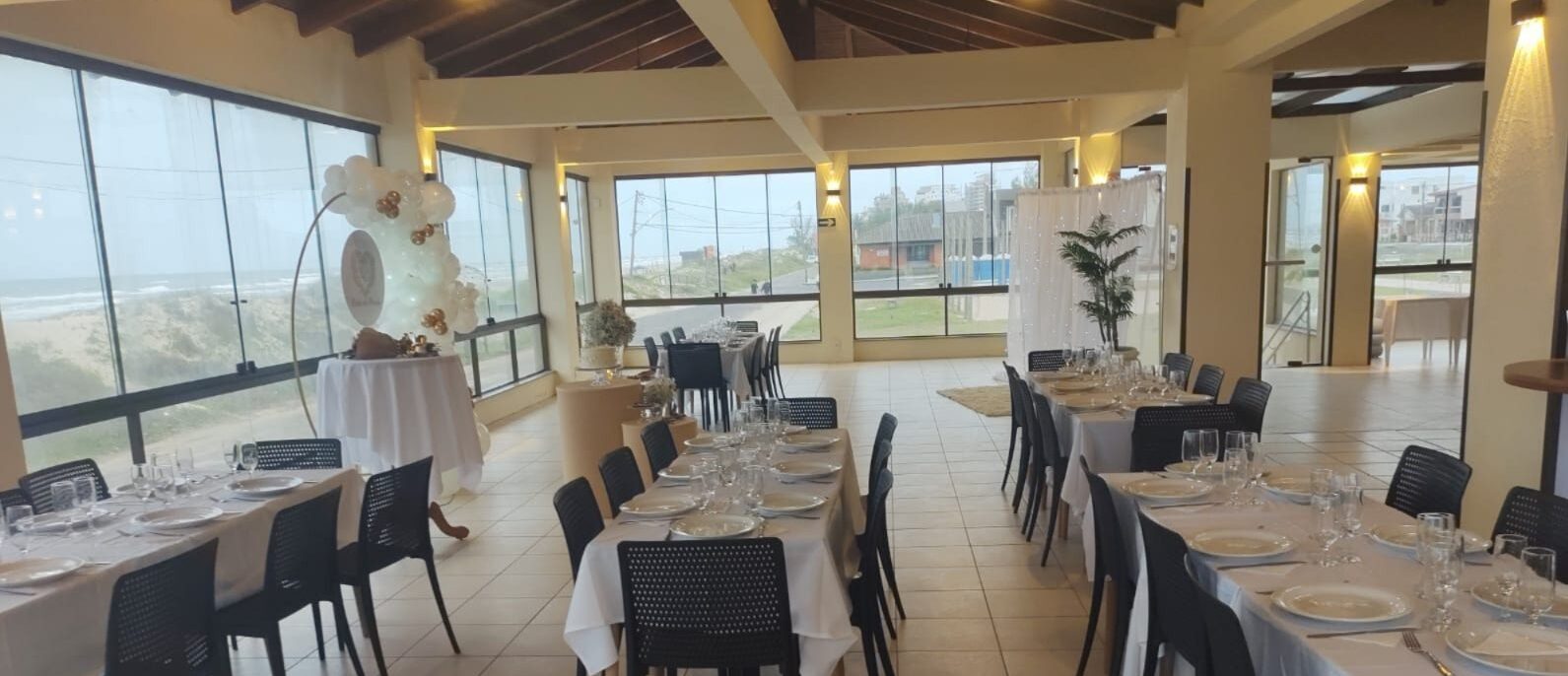 Salão de eventos com mesas compridas, cadeiras pretas, decoração floral branca, janelas amplas com vista para paisagem, iluminação moderna e teto com acabamento