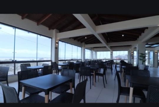 Restaurante moderno com mesas pretas, cadeiras cinzas e grandes janelas com vista para paisagem ao fundo, teto com vigas brancas