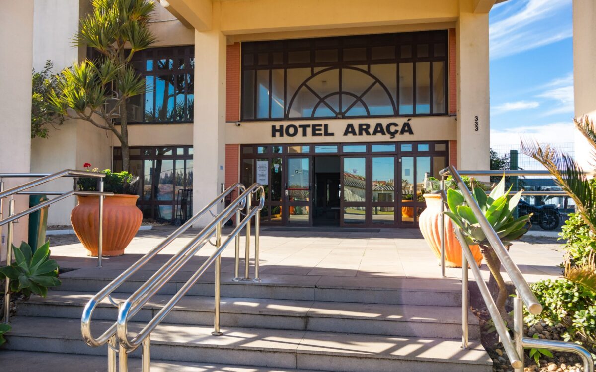 Fachada de hotel com entrada de escadas, rampas metálicas, vasos de cerâmica laranja, plantas e janelas com vidro, sob céu azul