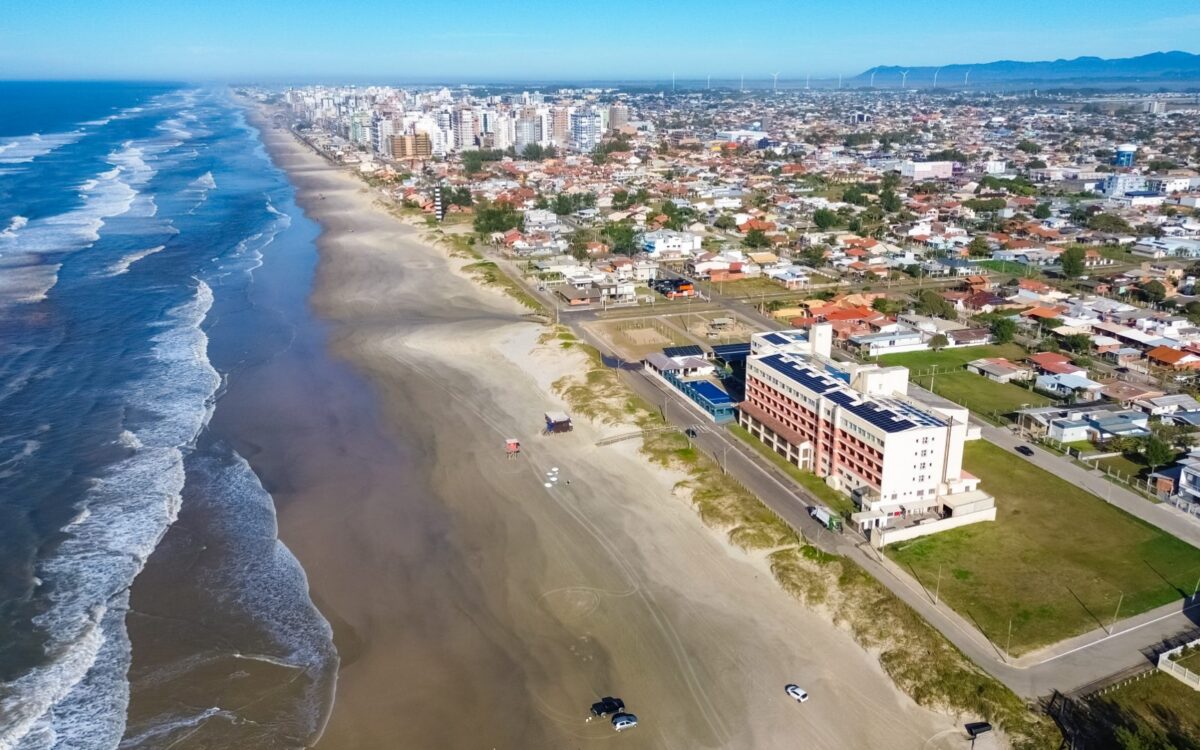 Vista aérea de praia com ondas, areia, edifícios à beira-mar, residências e montanhas ao fundo sob céu azul claro
