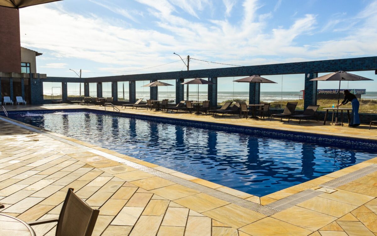 Piscina olímpica coberta com água azul, espreguiçadeiras, guarda-sóis, vista para o mar e praia ao fundo sob céu azul com nuvens