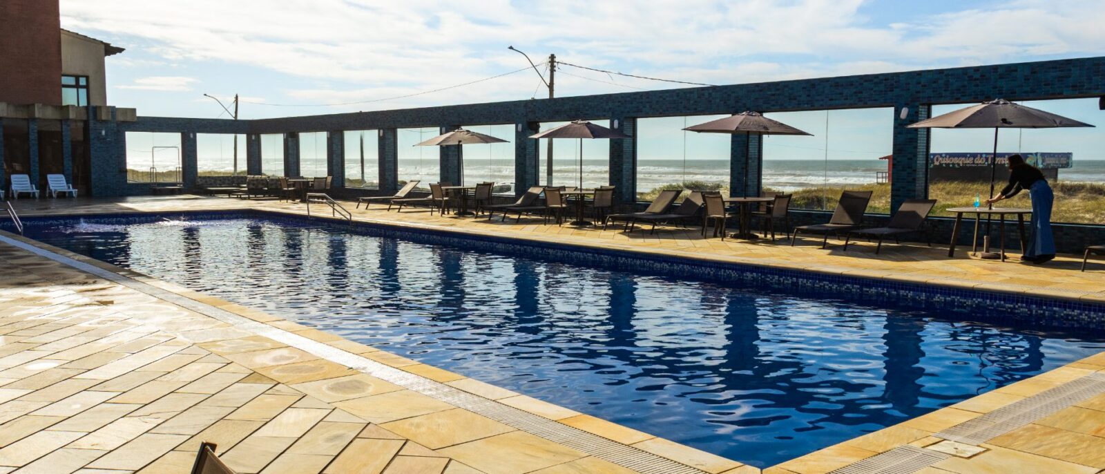 Piscina olímpica coberta com água azul, espreguiçadeiras, guarda-sóis, vista para o mar e praia ao fundo sob céu azul com nuvens