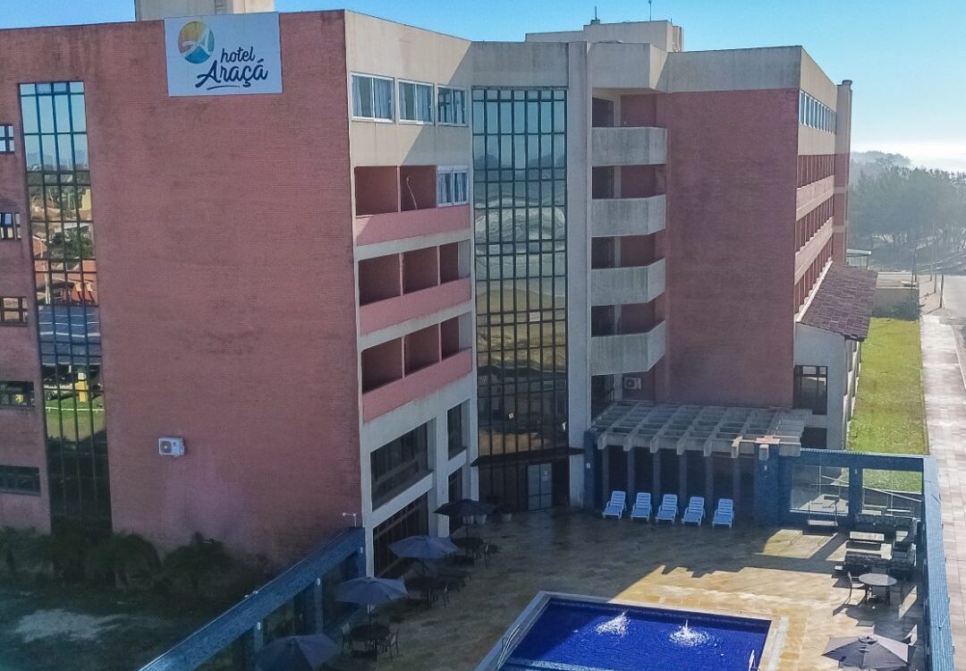 Vista aérea de um hotel com piscina retangular azul, espreguiçadeiras com guarda-sóis, edifícios de alvenaria vermelha e cinza, sob céu azul com nuvens