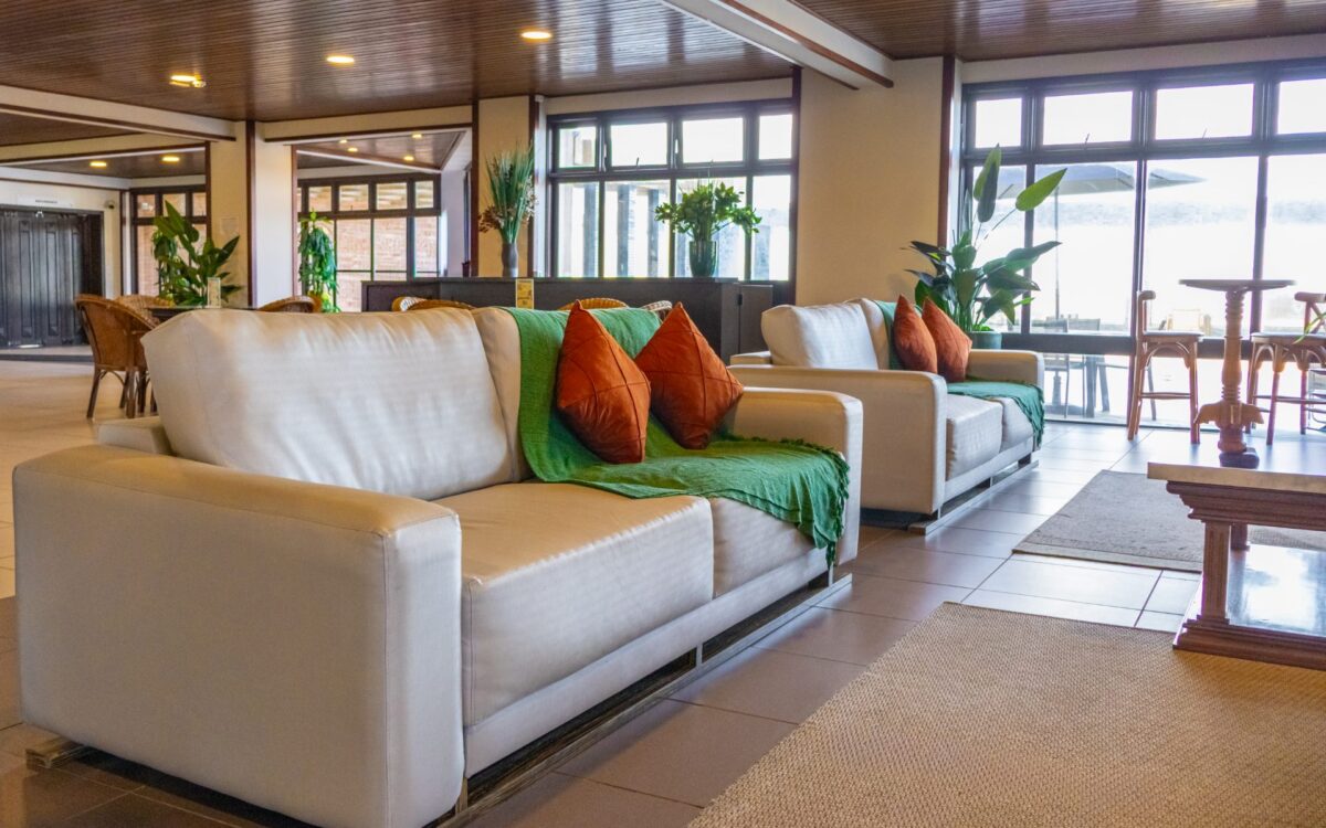 Sala de estar moderna com sofás brancos, almofadas coloridas em verde e laranja, plantas decorativas, grandes janelas com vista para o mar e teto de madeira