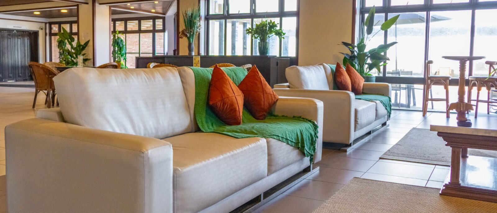 Sala de estar moderna com sofás brancos, almofadas coloridas em verde e laranja, plantas decorativas, grandes janelas com vista para o mar e teto de madeira