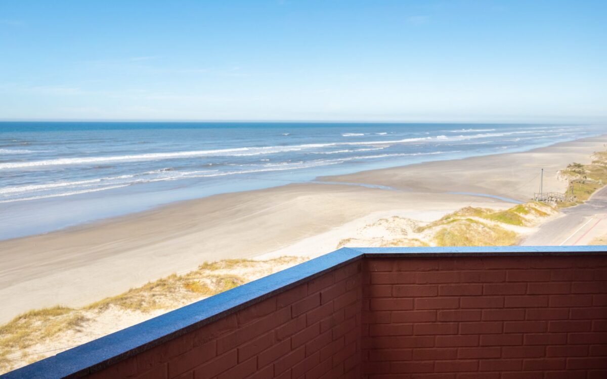 Vista de uma praia ampla com areia clara, ondas do mar e duna com vegetação, observada de uma estrutura com parede marrom e borda azul