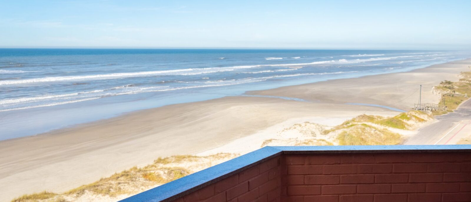 Vista de uma praia ampla com areia clara, ondas do mar e duna com vegetação, observada de uma estrutura com parede marrom e borda azul